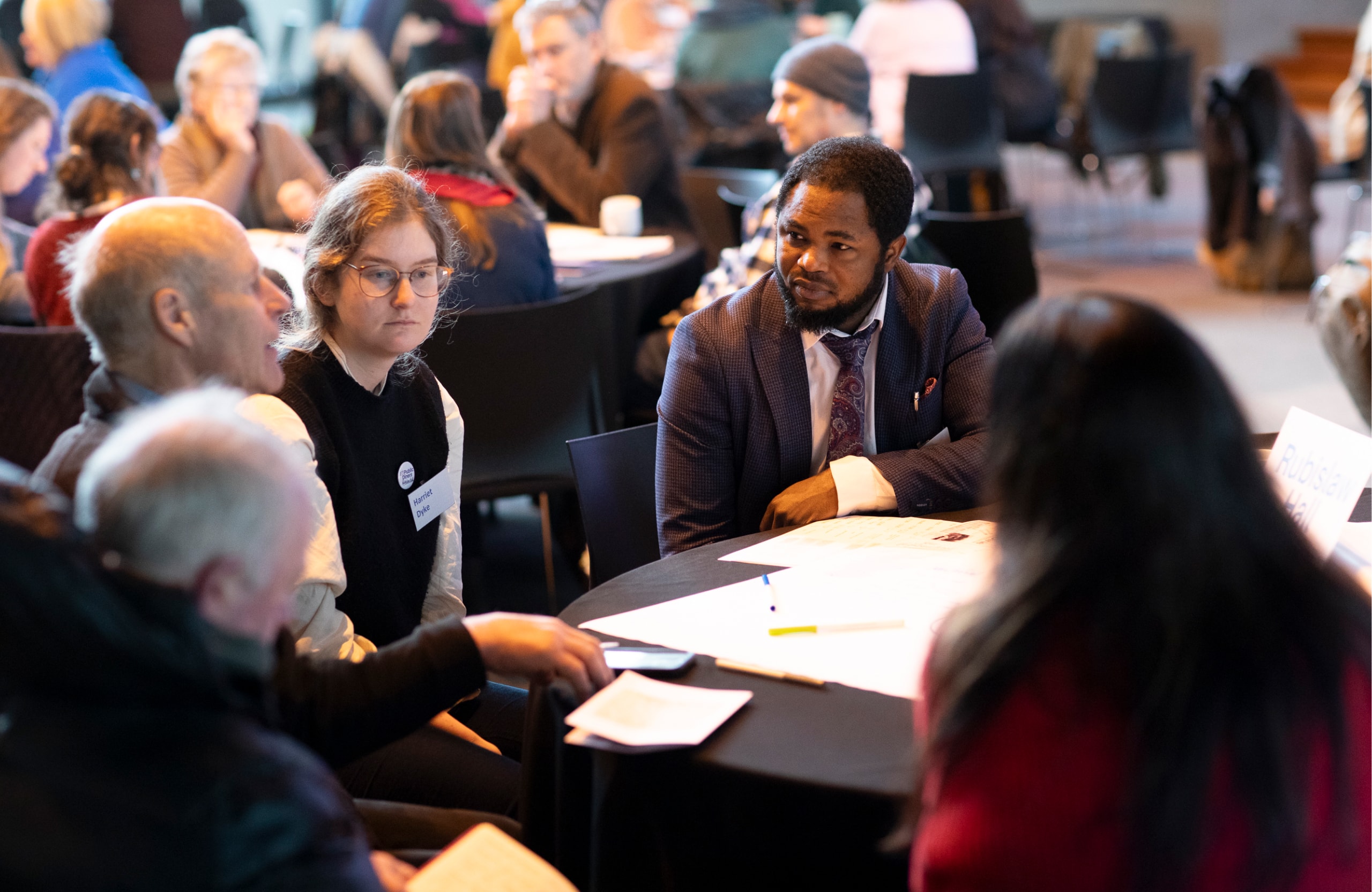 Group of people sit around a table  discussing a topic. People in the background doing the same.