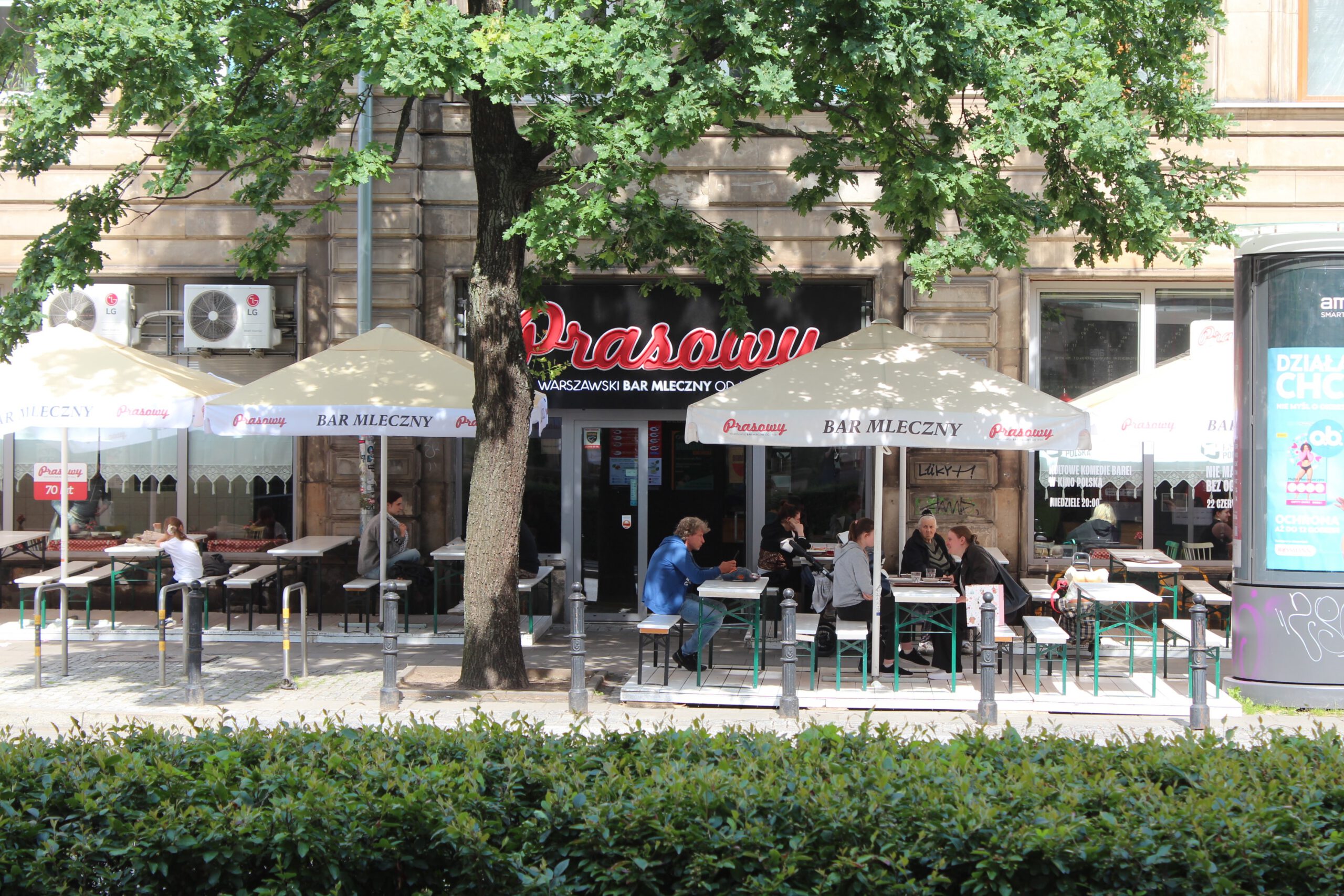 Outside view of Polish milk bar 'Prasowy', with diners seated at shaded tables under a mature oak tree.